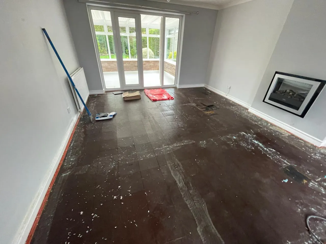 An empty room showing a dark, damaged subfloor with white debris and adhesive residue, consistent with asbestos floor tile removal.