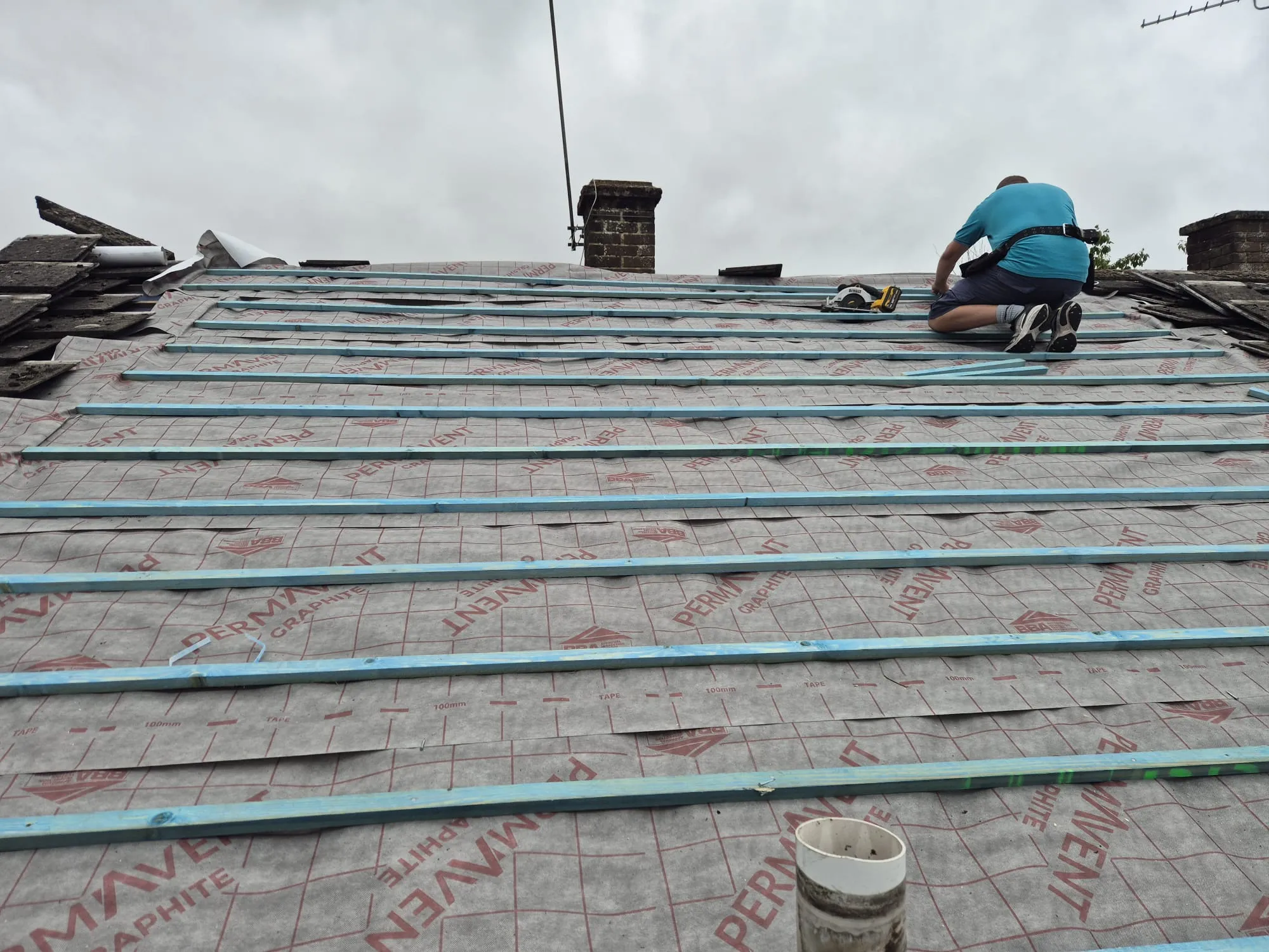 A roofer at work installing battens and underlayment on a pitched roof replacement project.