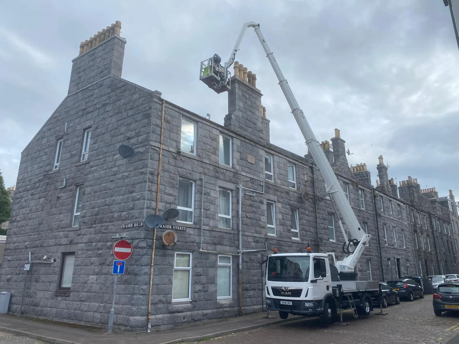 A worker in a cherry picker basket undertaking chimney repairs on a grey stone residential building.