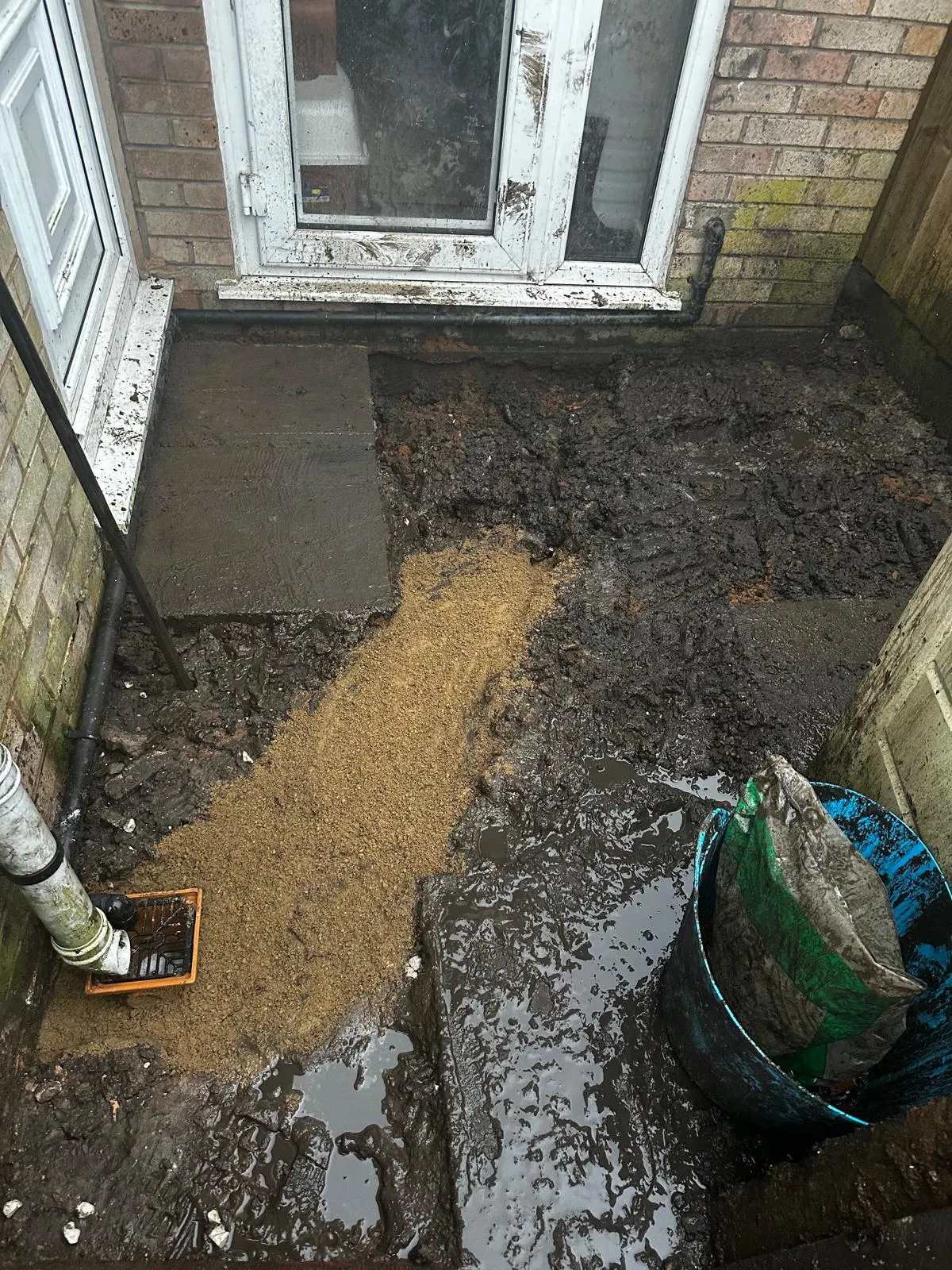 An image showing a construction site for drainage work outside a home, with excavated earth, mud, and a newly installed drain gulley and pipe.