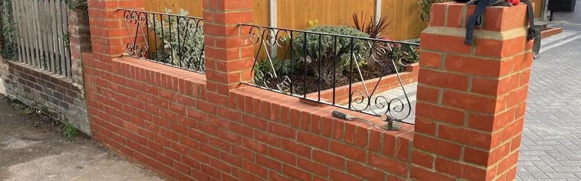 Newly constructed red brick garden wall with elegant black wrought iron railings and grey block paving.