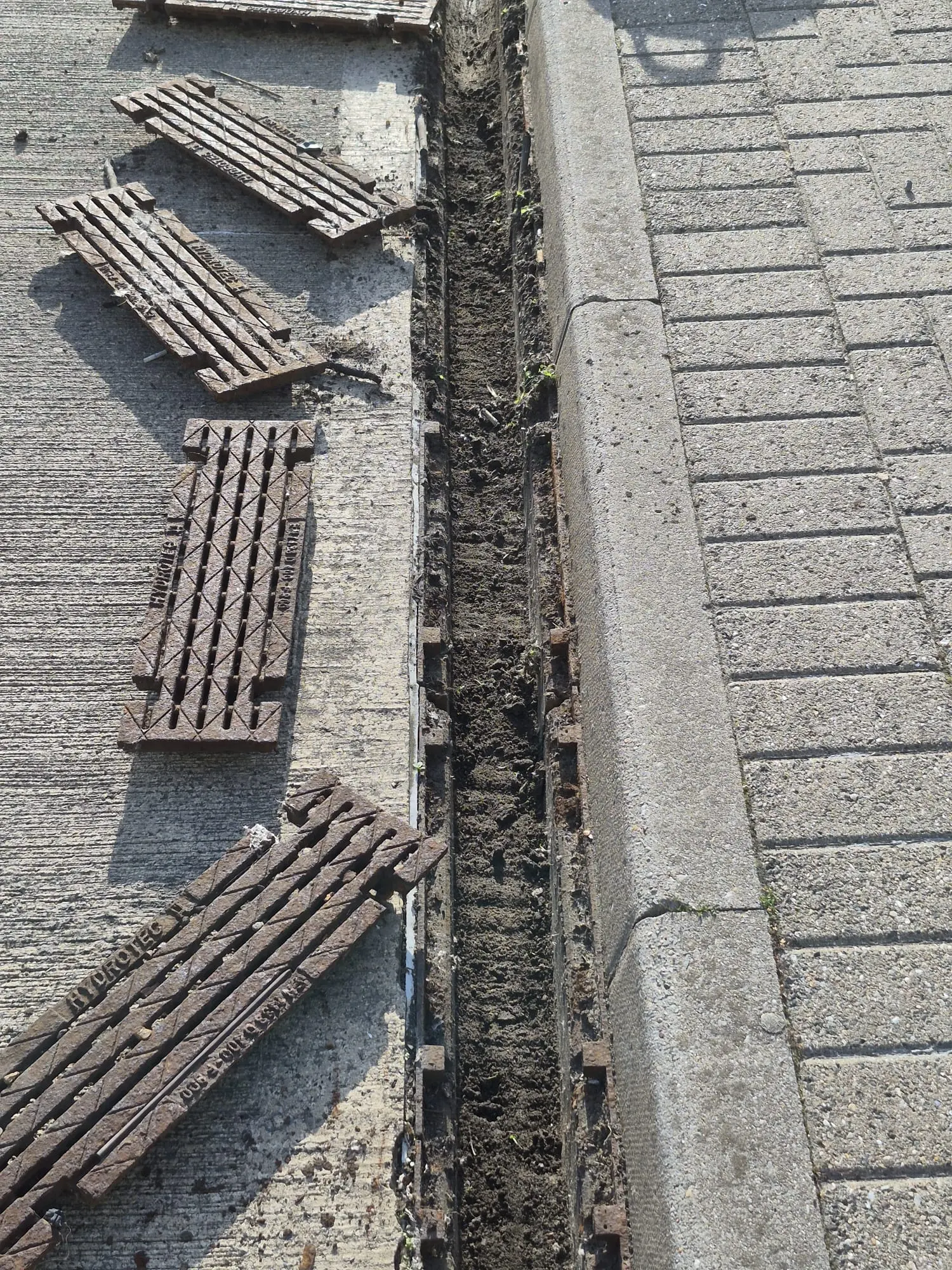 A heavily blocked commercial linear drainage channel with its cast iron grates removed, exposing a thick buildup of dirt and debris inside.