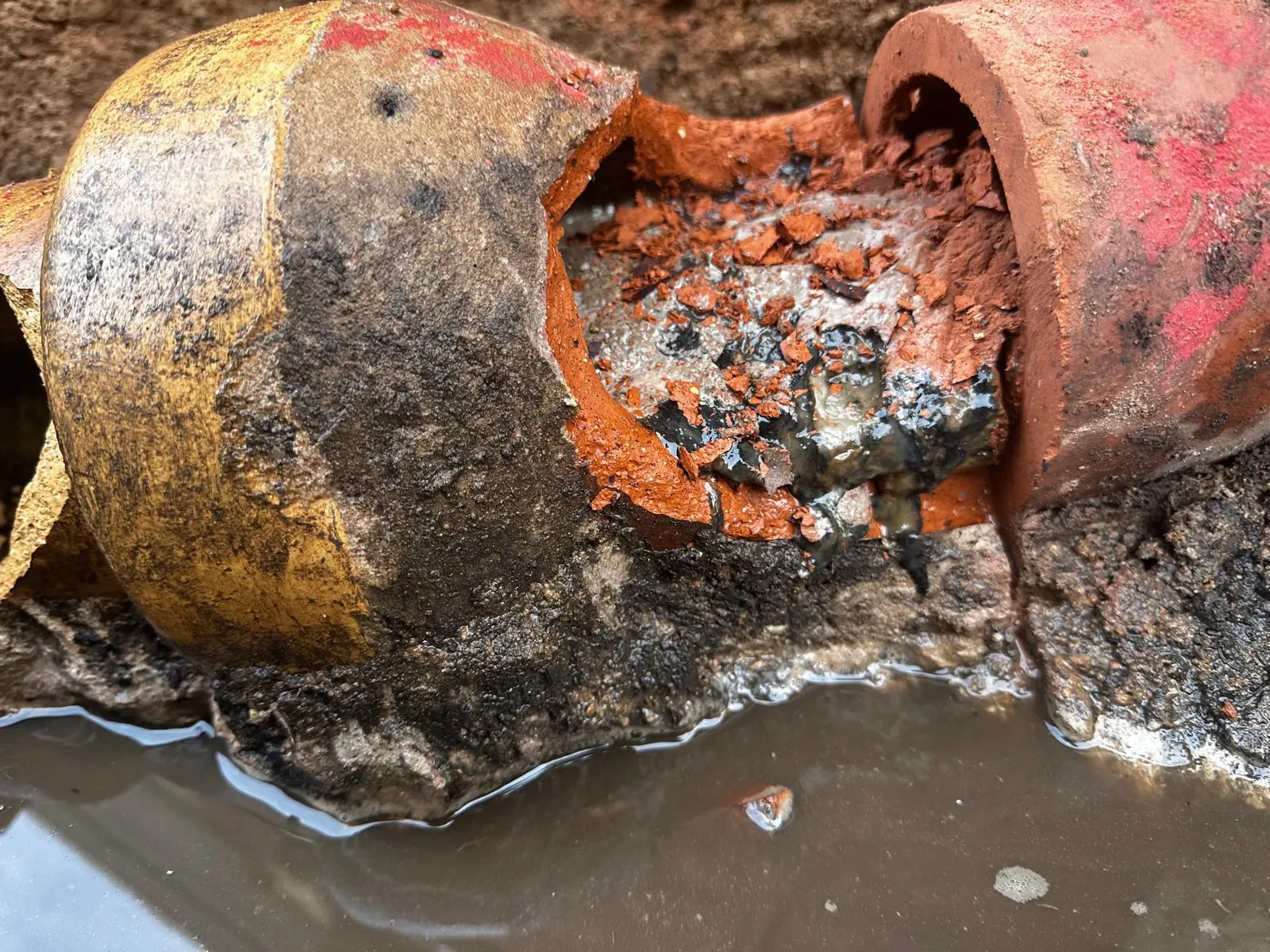 Close-up of a collapsed or broken clay drain pipe in an excavated area, revealing a blockage and standing wastewater, illustrating the need for drain repair.