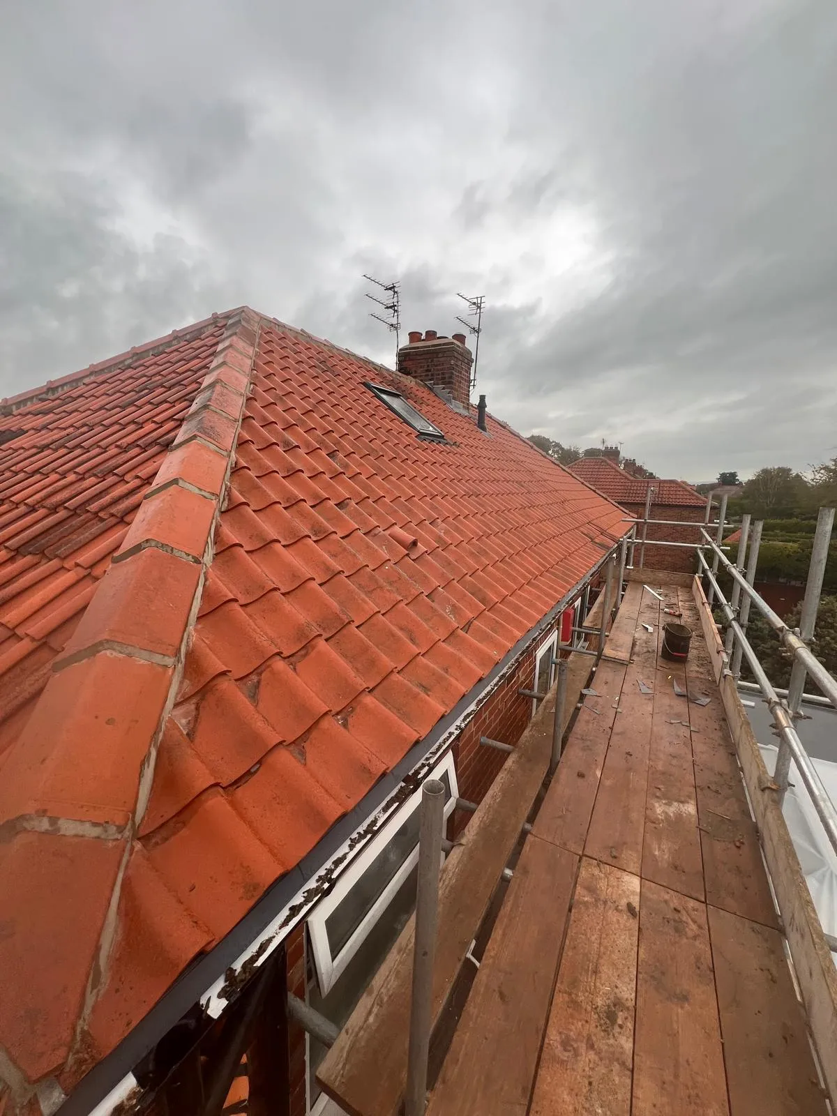 A close-up view of a residential terracotta tiled pitched roof with scaffolding, a chimney stack, and a small roof window under a cloudy sky.