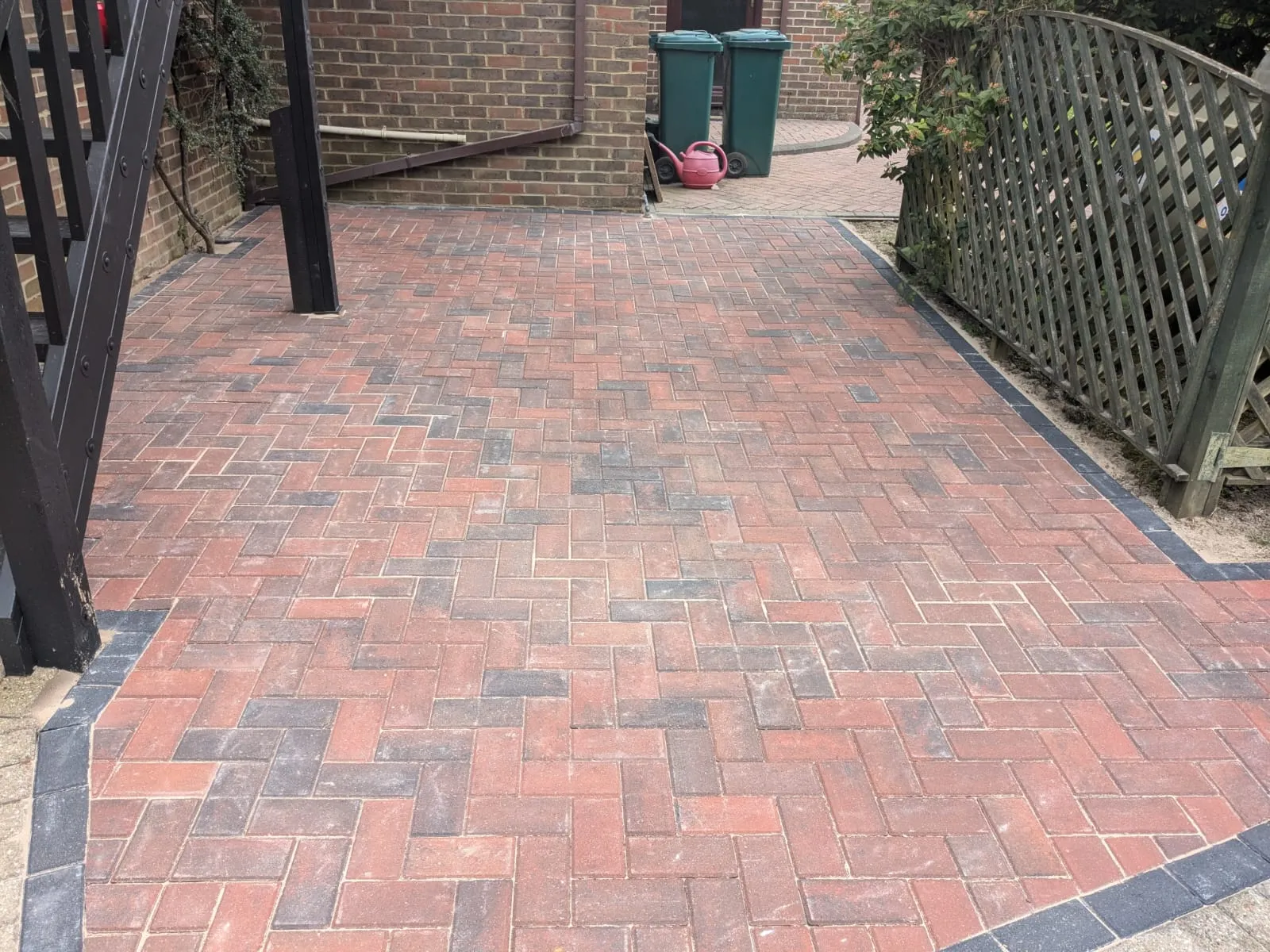 Newly laid block paving driveway or patio in red and charcoal herringbone pattern with a dark border, next to a brick house wall and wooden fence in a residential garden.