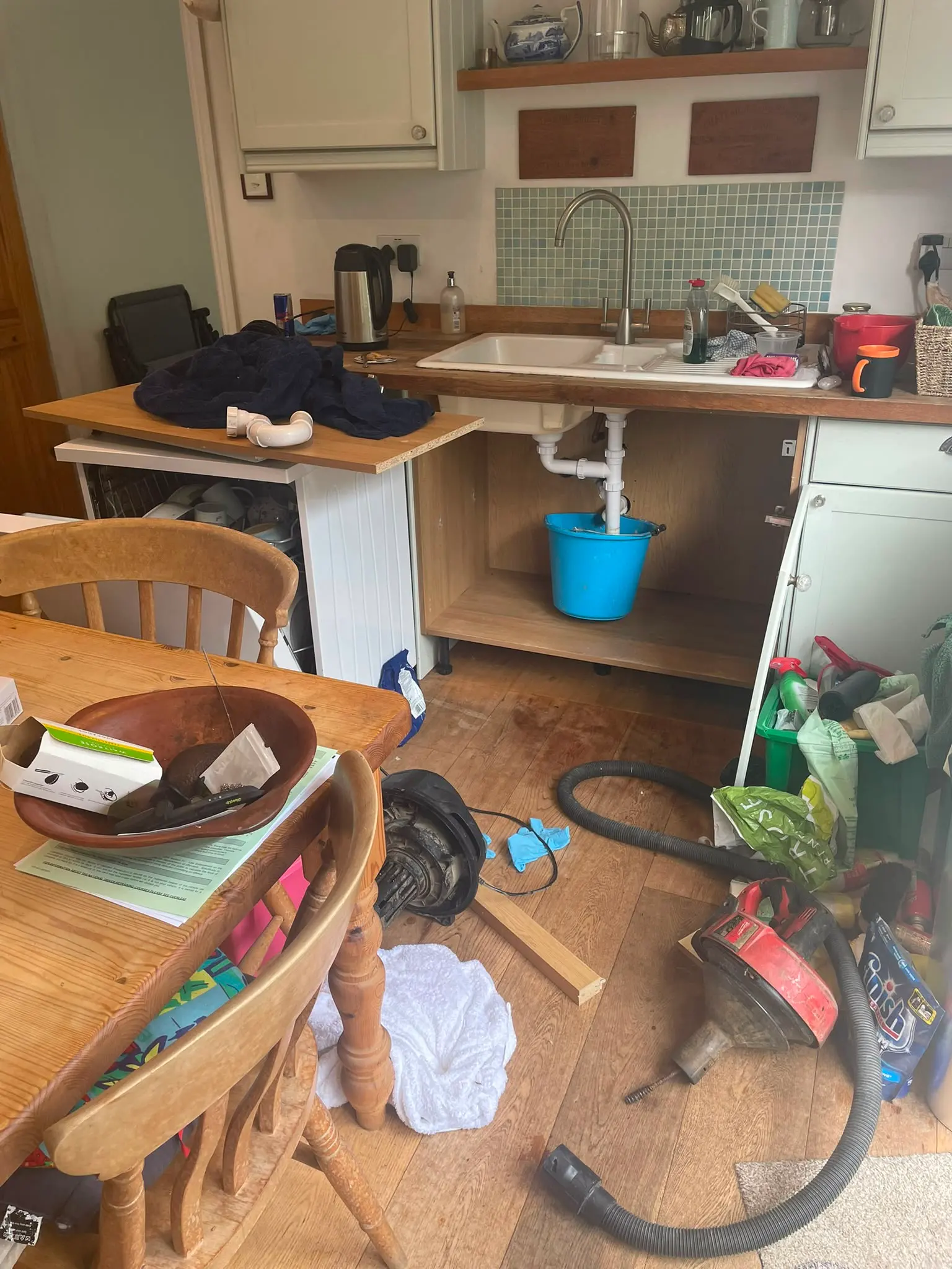 Kitchen sink plumbing repair in progress showing exposed pipes, a bucket, and professional drain unblocking tools on the floor.