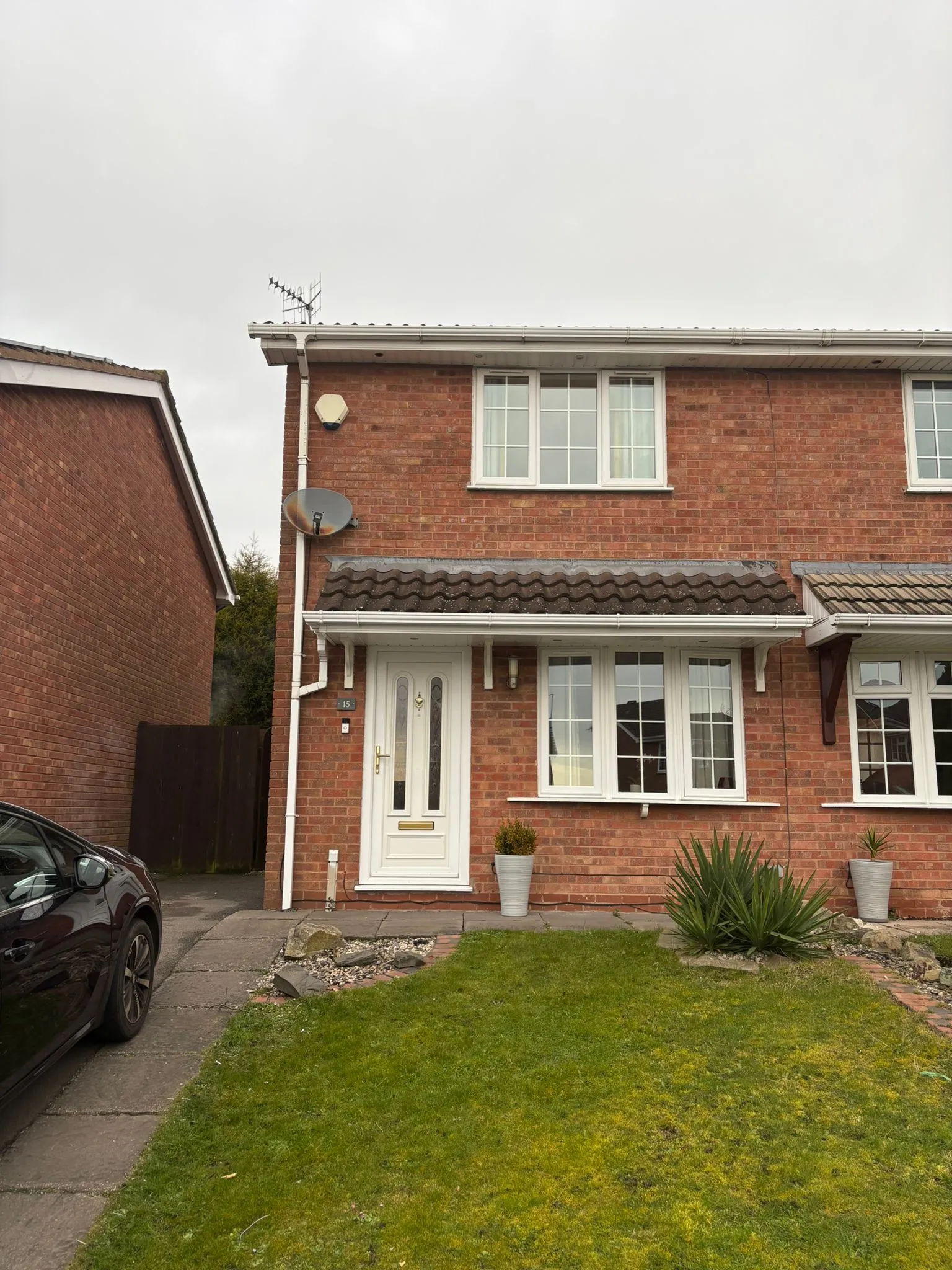 Front view of a red brick semi-detached house with white frames, a small lawn, and a car parked on the driveway, illustrating a property that could be extended.