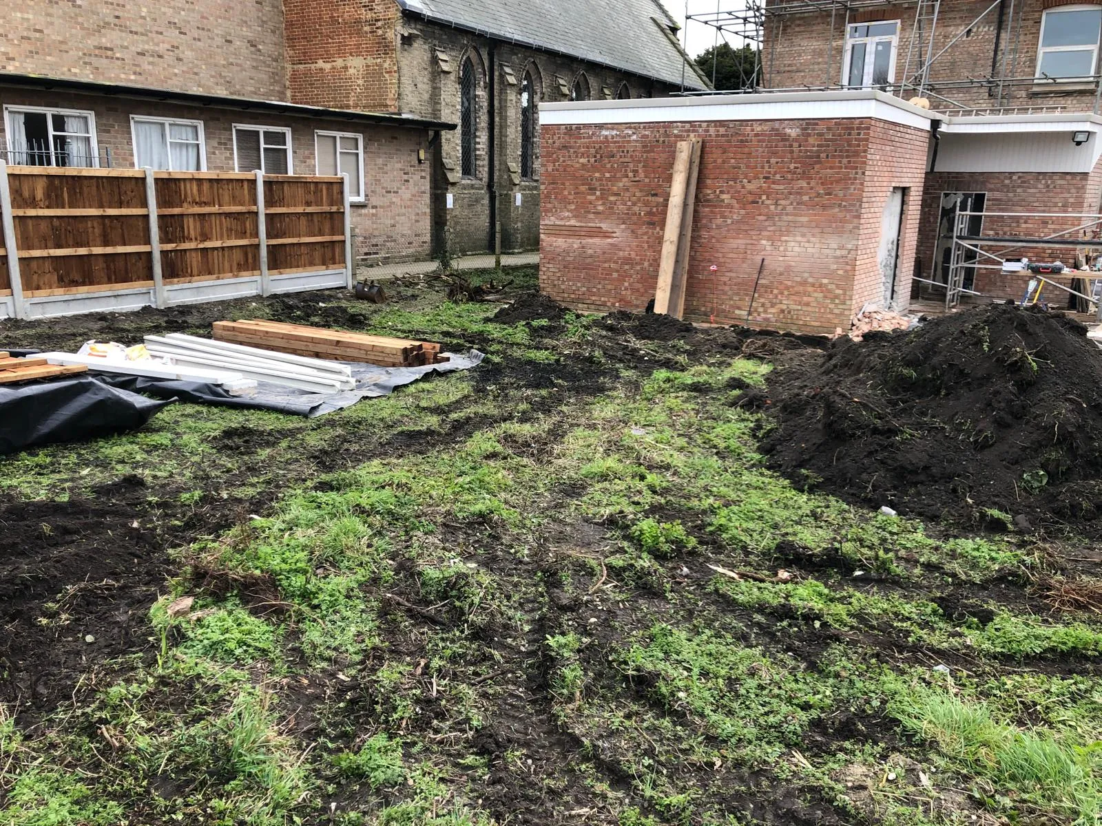 Construction site with a new brick extension, timber, new fencing, and muddy ground, indicating ongoing building and renovation work.