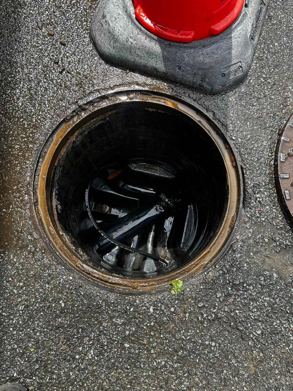 An open manhole showing a jet wash cleaning hose inside, indicating drain unblocking or inspection work.
