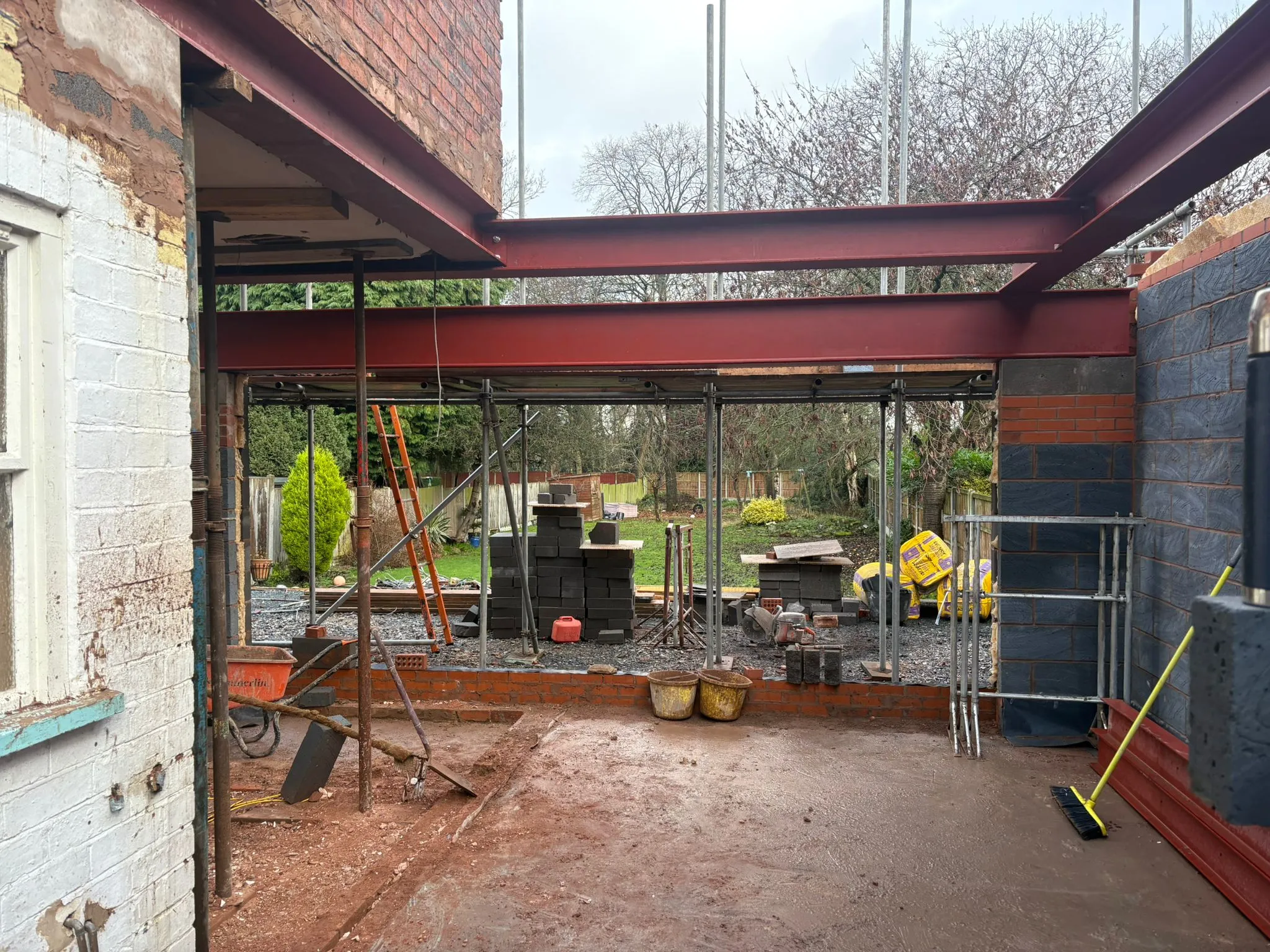 A construction site for a home extension, viewed from inside. Large red structural steel beams are in place, forming the upper framework. New grey and red brickwork is visible, alongside scaffolding, building materials like stacks of blocks, and tools. The ground is muddy, and a garden is visible through the open framework.