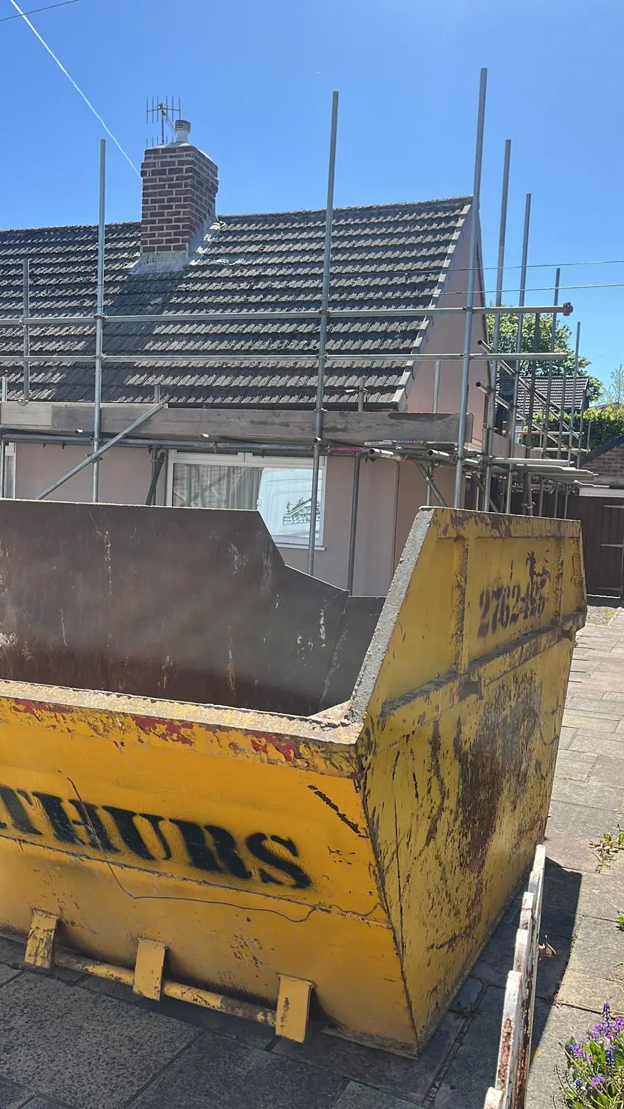 Metal scaffolding set up around a residential tiled roof with a large yellow skip in the driveway for a roof replacement project